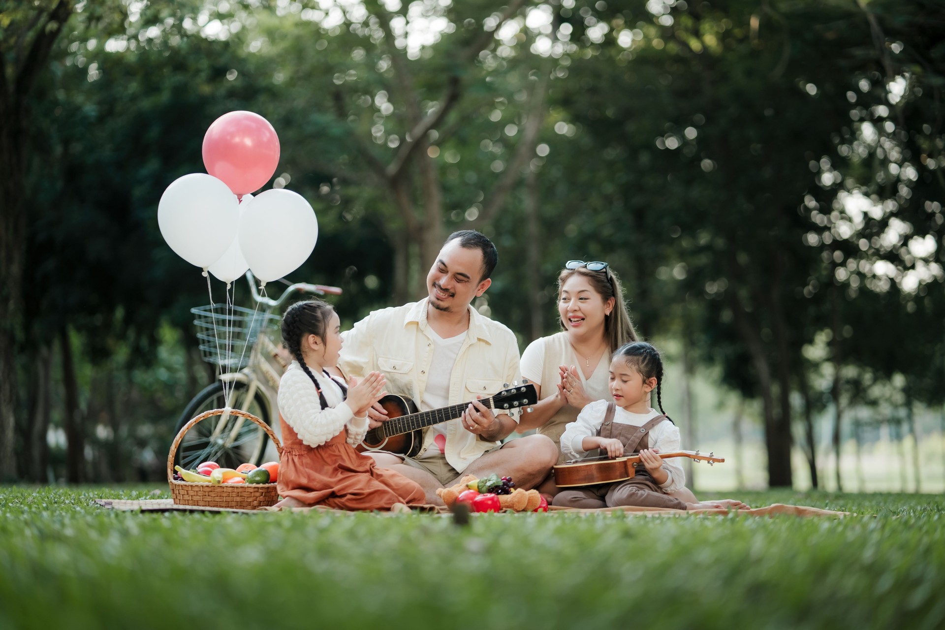 A family of four is sitting on a blanket in a park, with a man playing a guitar. The scene is joyful and relaxed, with the family enjoying each other's company and the music